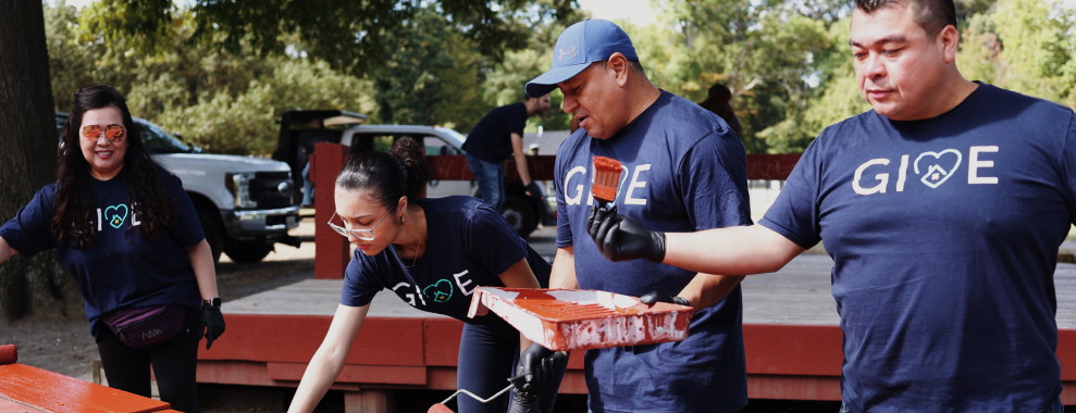 Four individuals painting wood, all wearing matching navy blue shirts with the GIVE logo and heart symbol